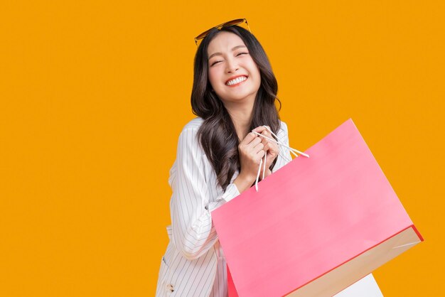 아시아의 행복한 여성 여자는 노란색 배경 스튜디오 샷에 다채로운 쇼핑 패키지를 들고 가방을 들고 카메라를 보며 웃고 있는 초상화를 닫습니다