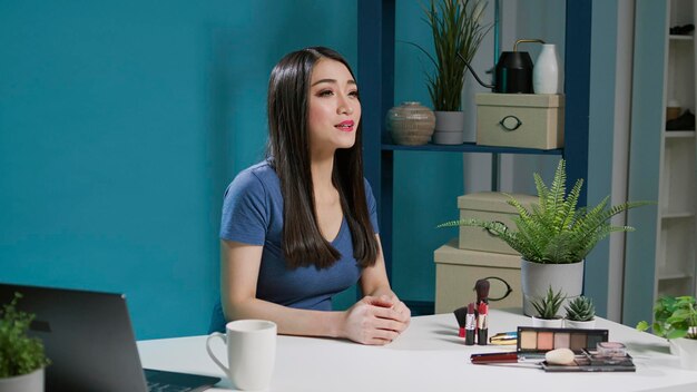 Asian influencer filming makeup tutorial video in studio, using smartphone camera to record. Content creator streaming videoblog on mobile phone with cosmetics, eyeshadow palette and lipstick.