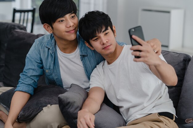 Asian influencer Gay couple vlog at home. Asian LGBTQ men happy relax fun using technology mobile phone record lifestyle vlog video upload in social media while lying sofa in living room .