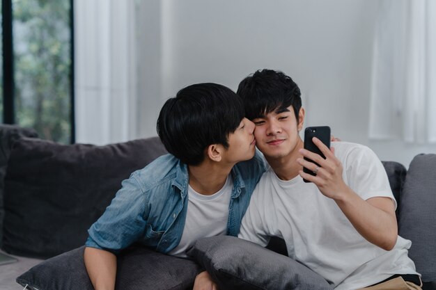 Asian influencer Gay couple vlog at home. Asian LGBTQ men happy relax fun using technology mobile phone record lifestyle vlog video upload in social media while lying sofa in living room .