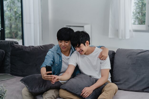 집에서 아시아 영향 게이 커플 블로그. 아시아 LGBTQ 남성은 거실에 소파에 누워있는 동안 소셜 미디어에 기술 휴대 전화 기록 라이프 스타일 vlog 비디오 업로드를 사용하여 즐거운 휴식을 취하십시오.