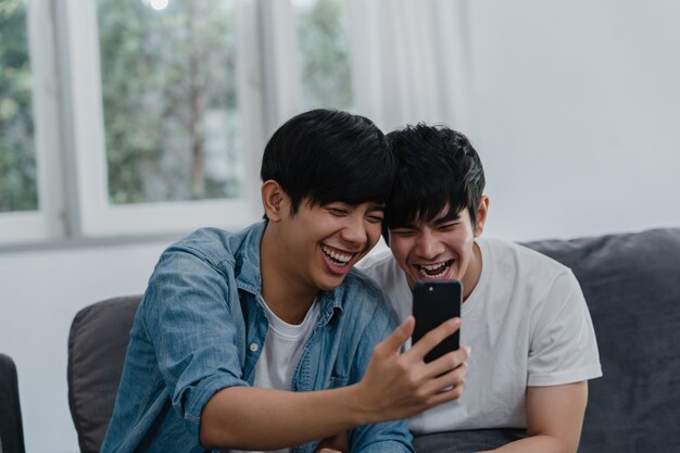 Asian influencer Gay couple vlog at home. Asian LGBTQ men happy relax fun using technology mobile phone record lifestyle vlog video upload in social media while lying sofa in living room .