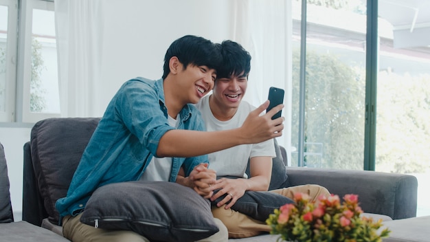 Asian influencer Gay couple vlog at home. Asian LGBTQ+ men happy relax fun using technology mobile phone record lifestyle vlog video upload in social media while lying sofa in living room .