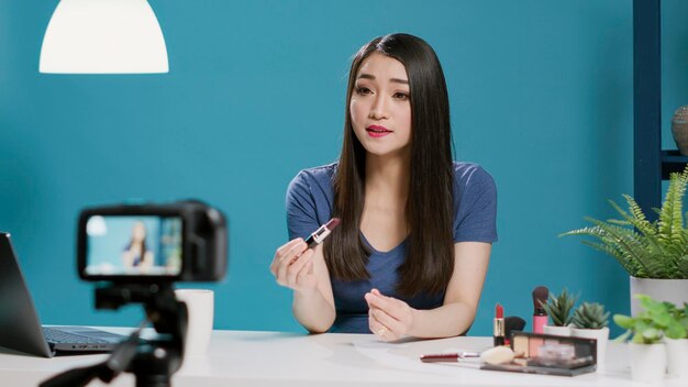 Asian influencer reviewing lipstick product on vlogging camera, filming recommendations video with makeup cosmetics. Young millennial vlogger broadcasting discussion on online internet.