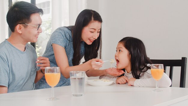 Asian Japanese family has breakfast at home. Asian happy dad, mom, and daughter eat spaghetti drink orange juice on table in modern kitchen at house in the morning .