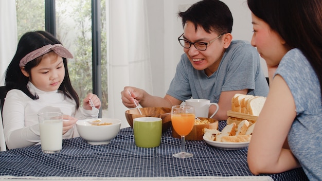 アジア系の日本人家族は家で朝食をとります。アジアのママ、パパ、娘は、朝、台所のテーブルの上のボウルでパン、コーンフレーク、シリアル、ミルクを食べながら一緒に話して幸せを感じています。