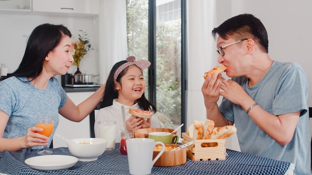 アジア系の日本人家族は家で朝食をとります。アジアのママ、パパ、娘は、朝、台所のテーブルの上のボウルでパン、コーンフレーク、シリアル、ミルクを食べながら一緒に話して幸せを感じています。