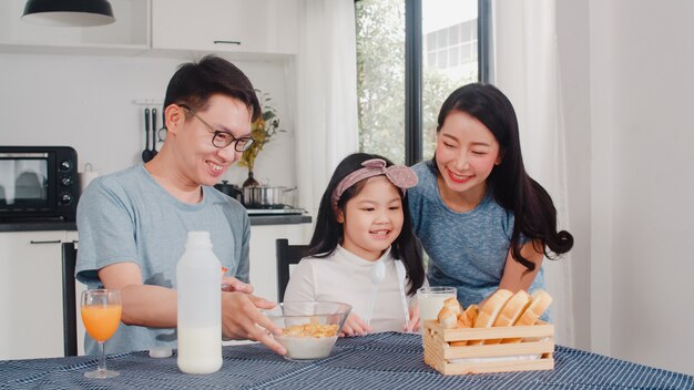 アジア系の日本人家族は家で朝食をとります。アジアのママ、パパ、娘は、朝、台所のテーブルの上のボウルでパン、コーンフレーク、シリアル、ミルクを食べながら一緒に話して幸せを感じています。