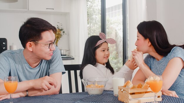 アジア系の日本人家族は家で朝食をとります。アジアのママ、パパ、娘は、朝、台所のテーブルの上のボウルでパン、コーンフレーク、シリアル、ミルクを食べながら一緒に話して幸せを感じています。