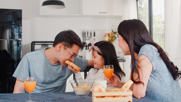 Asian Japanese family has breakfast at home. Asian mom, dad, and daughter feeling happy talking together while eat bread, corn flakes cereal and milk in bowl on table in the kitchen in the morning.