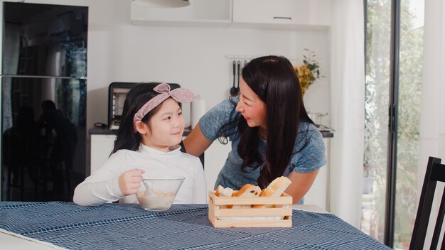 アジア系の日本人家族は家で朝食をとります。アジアのお母さんと娘は、朝の家でモダンなキッチンのテーブルの上のボウルにパン、コーンフレークシリアル、牛乳を食べながら一緒に話して幸せを感じます。