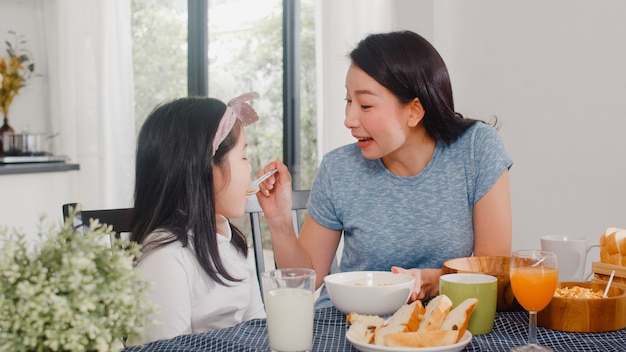 アジア系の日本人家族は家で朝食をとります。アジアの母と娘が一緒に幸せな話をしながらパンを食べて、オレンジジュース、コーンフレークシリアル、牛乳をモダンなキッチンのテーブルの上の朝に飲みます。