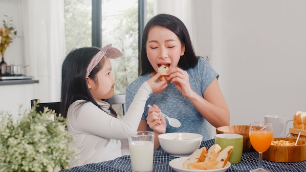 Asian Japanese family has breakfast at home. Asian mom and daughter happy talking together while eating bread, drink orange juice, corn flakes cereal and milk on table in modern kitchen in morning.