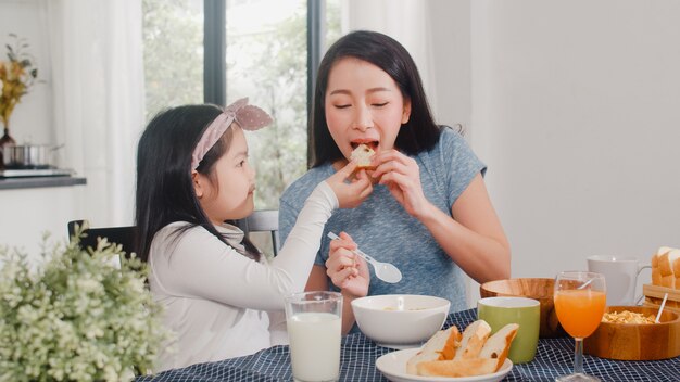 Asian Japanese family has breakfast at home. Asian mom and daughter happy talking together while eating bread, drink orange juice, corn flakes cereal and milk on table in modern kitchen in morning.