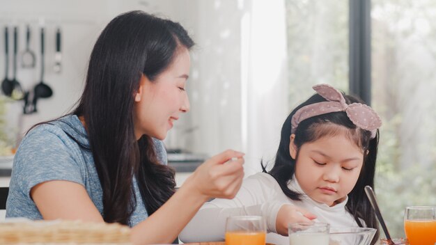 アジア系の日本人家族は家で朝食をとります。アジアの母と娘が一緒に幸せな話をしながらパンを食べて、オレンジジュース、コーンフレークシリアル、牛乳をモダンなキッチンのテーブルの上の朝に飲みます。