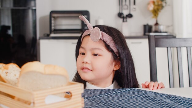 アジアの日本人の女の子は家でパンを食べる。朝は家のモダンなキッチンのテーブルの上のテーブルにそれを置く幸せなピックサンドイッチを感じているアジアの女性。