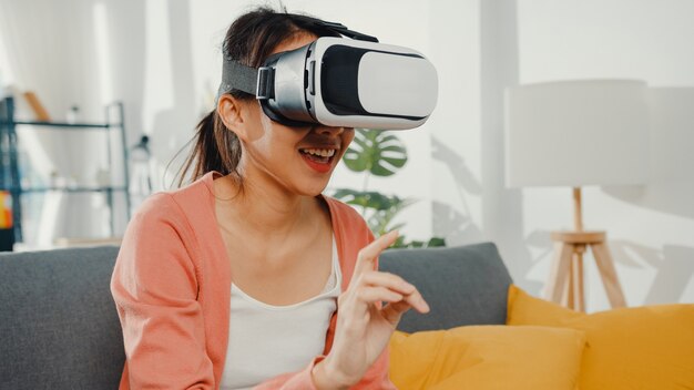asian lady wearing headset glasses of virtual reality gesticulating hand sitting on couch in living room at house.