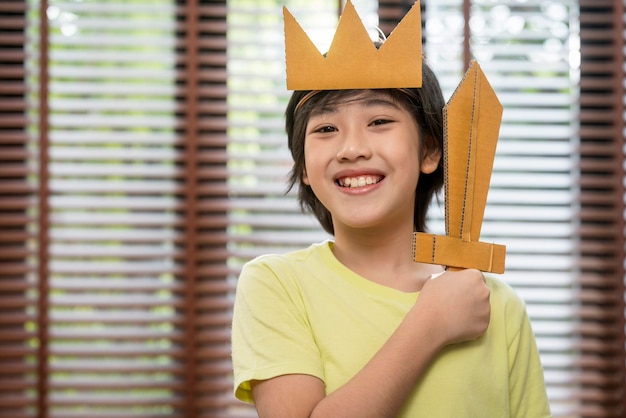 アジアの小さな王子の子の男の子の役割を演じる王の手として遊ぶ若い男の子は、陽気で楽しい紙の段ボールのゲームで笑顔の紙の剣を持っています