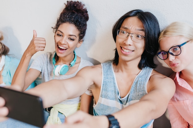 Free photo asian long-haired boy making selfie with friends holding black phone and smiling. funny blonde woman in glasses having fun with curly young lady and brunette student.