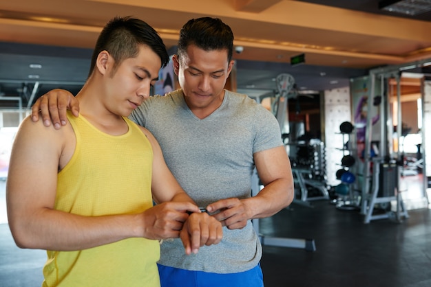 Asian male gym instructor with hand on client's shoulder looking at his fitness tracker 