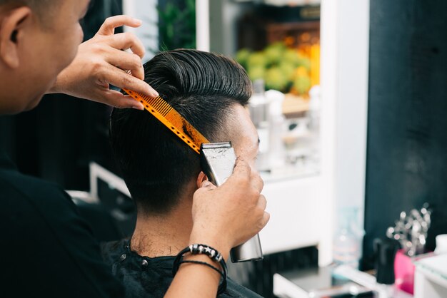 Азиатский мужской парикмахер держит расческу и триммер к голове клиента