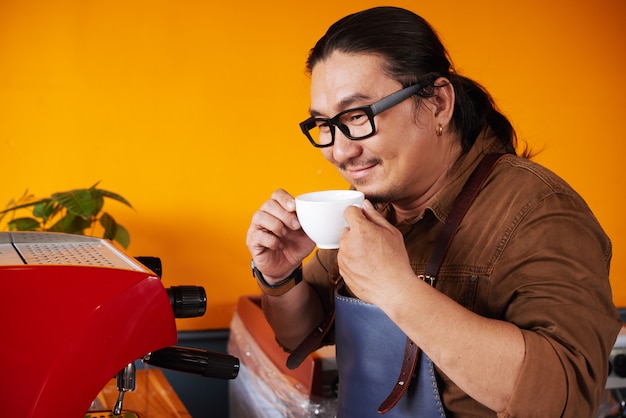 Asian man in apron standing next to espresso machine, holding cup and smelling coffee