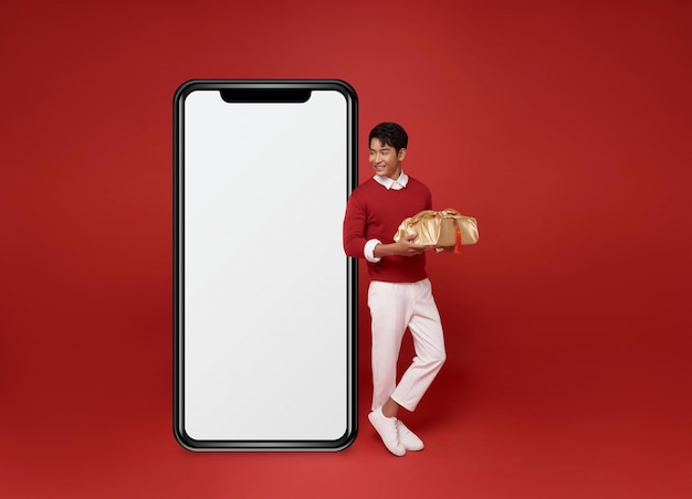 アジア系男性赤いスウェットを着てプレゼント箱を握って大きな空のスマートフォンの画面を見ています