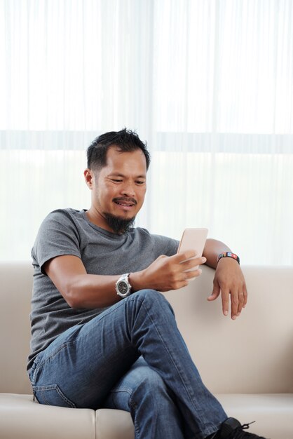 Азиатский человек неторопливо сидит на диване со скрещенными ногами и с помощью смартфона