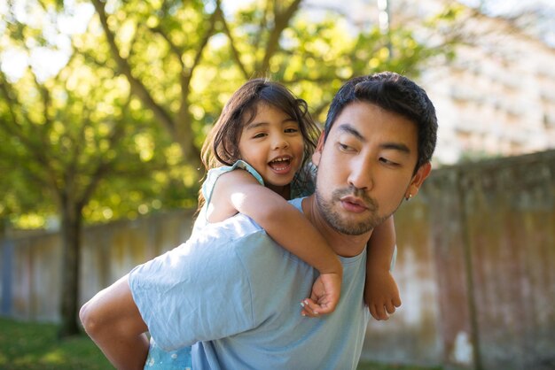 Азиатский мужчина и маленькая девочка веселятся в летнем парке. Радостный отец держит улыбающуюся дочь на спине, гуляя в парке, проводя выходные вместе. Детство, досуг, концепция летнего отдыха
