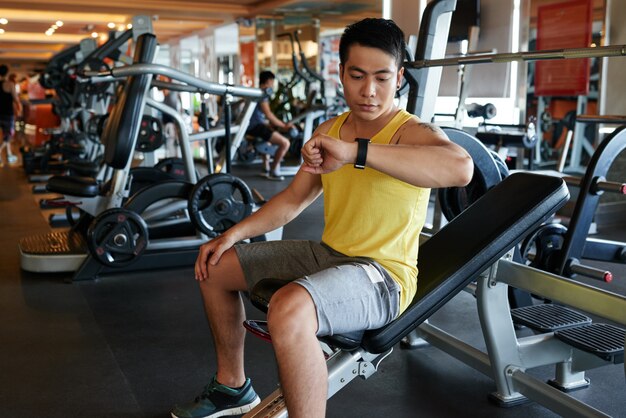 Asian man sitting on bench in gym and looking at wristwatch