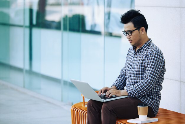 Asian man sitting on bench outdoors near glass wall and working on laptop