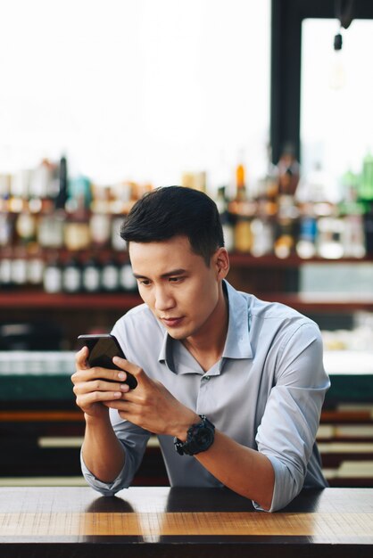 Asian man standing behind counter in bar and using smartphone