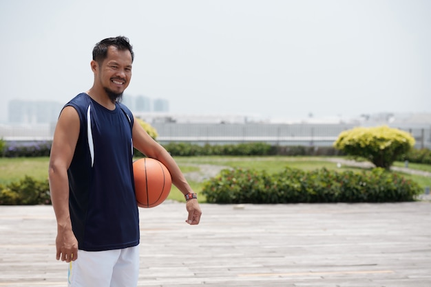 Foto gratuita uomo asiatico che sta all'aperto allo stadio, tenendo pallacanestro e sorridere