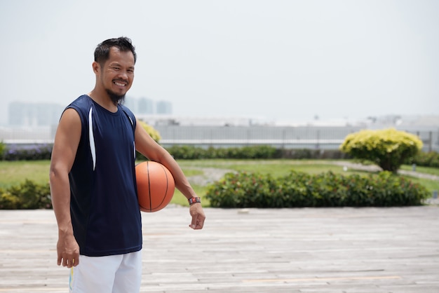 Asian man standing outdoors at stadium, holding basketball and smiling