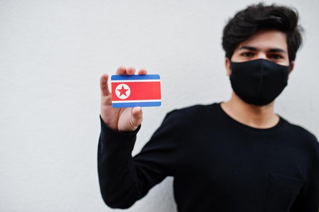 Азиатский мужчина носит все черное с маской для лица, держит в руке флаг Северной Кореи на белом фоне Коронавирусная концепция страны