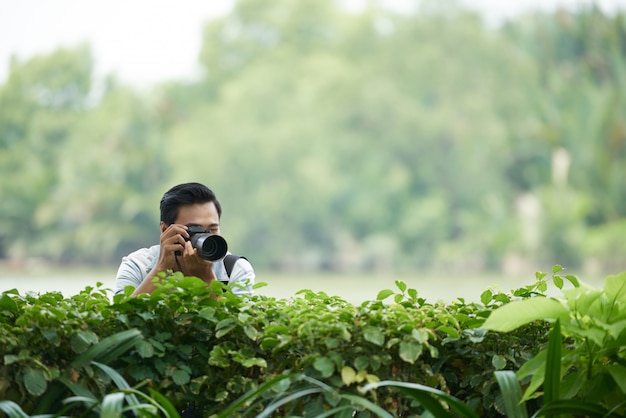 전문 카메라 공원에서 녹색 울타리를 통해 들여다 보며 사진을 찍는 아시아 사람
