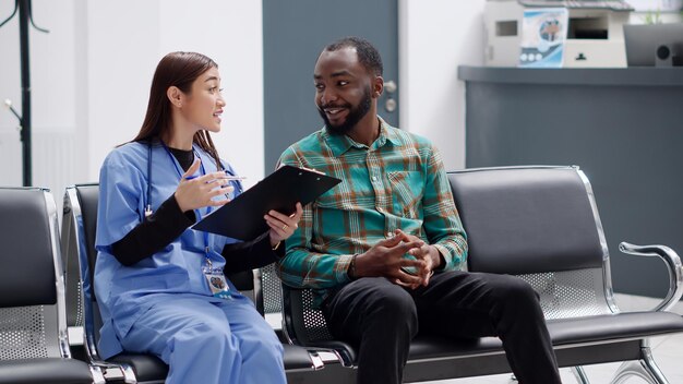 Asian medical assistant consulting male patient in medical clinic reception, sitting in waiting room lobby. Young man and nurse discussing about healthcare consultation and diagnosis.