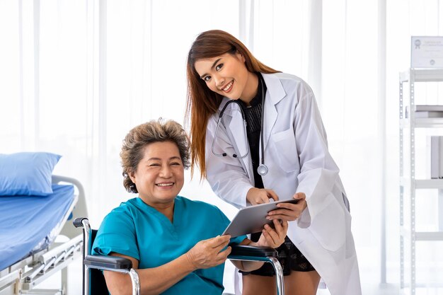 Asian medical doctor take care and explain senior elderly woman female patient on wheelchair with tablet looking at camera