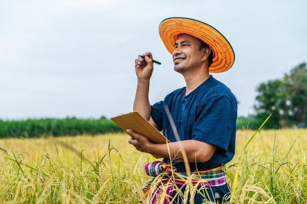 Asian middle aged farmer man wearing straw hat write on clipboard in rice field with smile during keep data