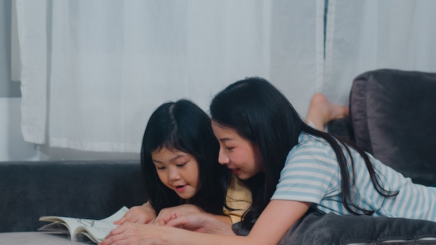 아시아 중년 여성이 그녀의 딸이 책을 읽고 자유 시간 집에서 휴식을 즐길 수 가르쳐. 라이프 스타일 어머니와 아이 행복 재미는 저녁에 현대 가정에서 거실에서 함께 시간을 보내십시오.