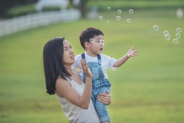 Asian mother and son blowing bubbles outdoor. cute toddler boy playing with soap bubbles on summer field. Hands up. happy childhood concept. authentic lifestyle image.