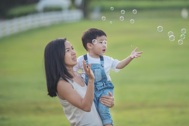 アジアの母と息子が屋外で泡を吹いています。夏の畑でシャボン玉で遊ぶかわいい幼児の男の子。手を挙げろ。幸せな子供時代のコンセプト。本格的なライフスタイルイメージ。