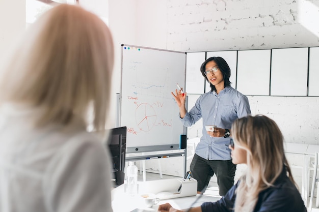 Free photo asian office manager writing on flipchart with colorful markers. indoor portrait of young blonde ladies watching chinese male colleague during his presentation.