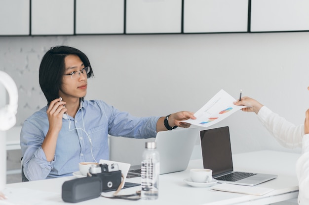 Asian office worker handed documents to colleague. Indoor portrait of brunette freelance programmer in earphones sitting with laptop and camera, while drinking coffee.