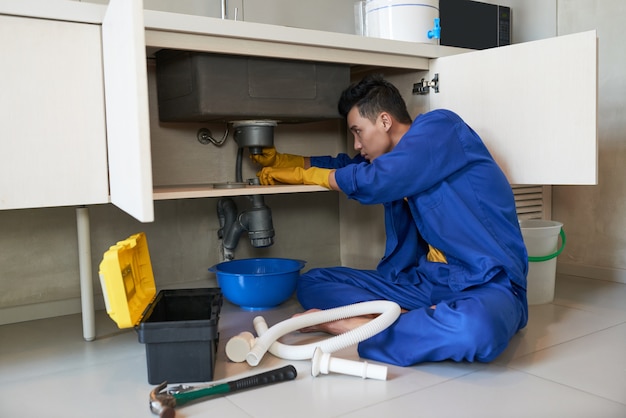 Free photo asian plumber in blue overalls clearing blockage in drain