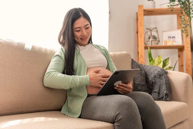 asian pregnant woman hands fondle on belly and holding tablet sitting on sofa at home doctor online