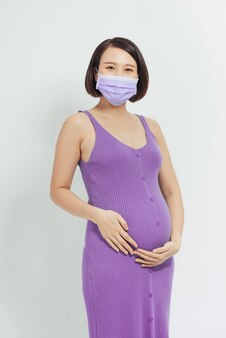 Asian pregnant woman wear face mask holding her belly, look at camera, isolated on white  background, covid-19 concept