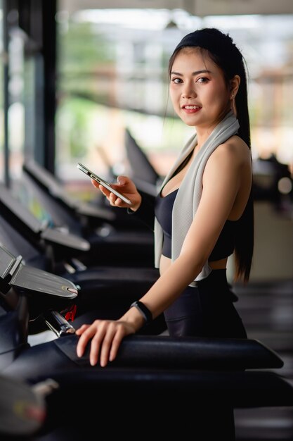 디딜 방아에 운동복과 smartwatch 나머지를 입고 아시아 예쁜 여자는 스마트 폰과 스마트 워치 운동 앱을 사용하고 현대 체육관에서 음악을 듣고