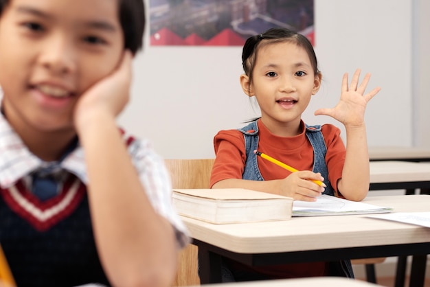 Asian pupils sitting in classroom and girl putting up hand to answer