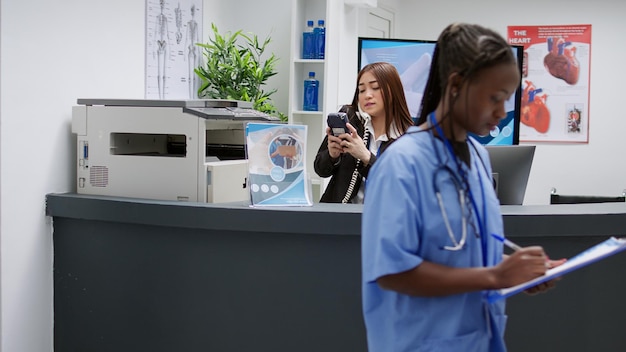 Asian receptionist answering landline phone call to help with checkup visit behind reception counter at medical facility. Clinic employee working in healthcare service, exam appointment.